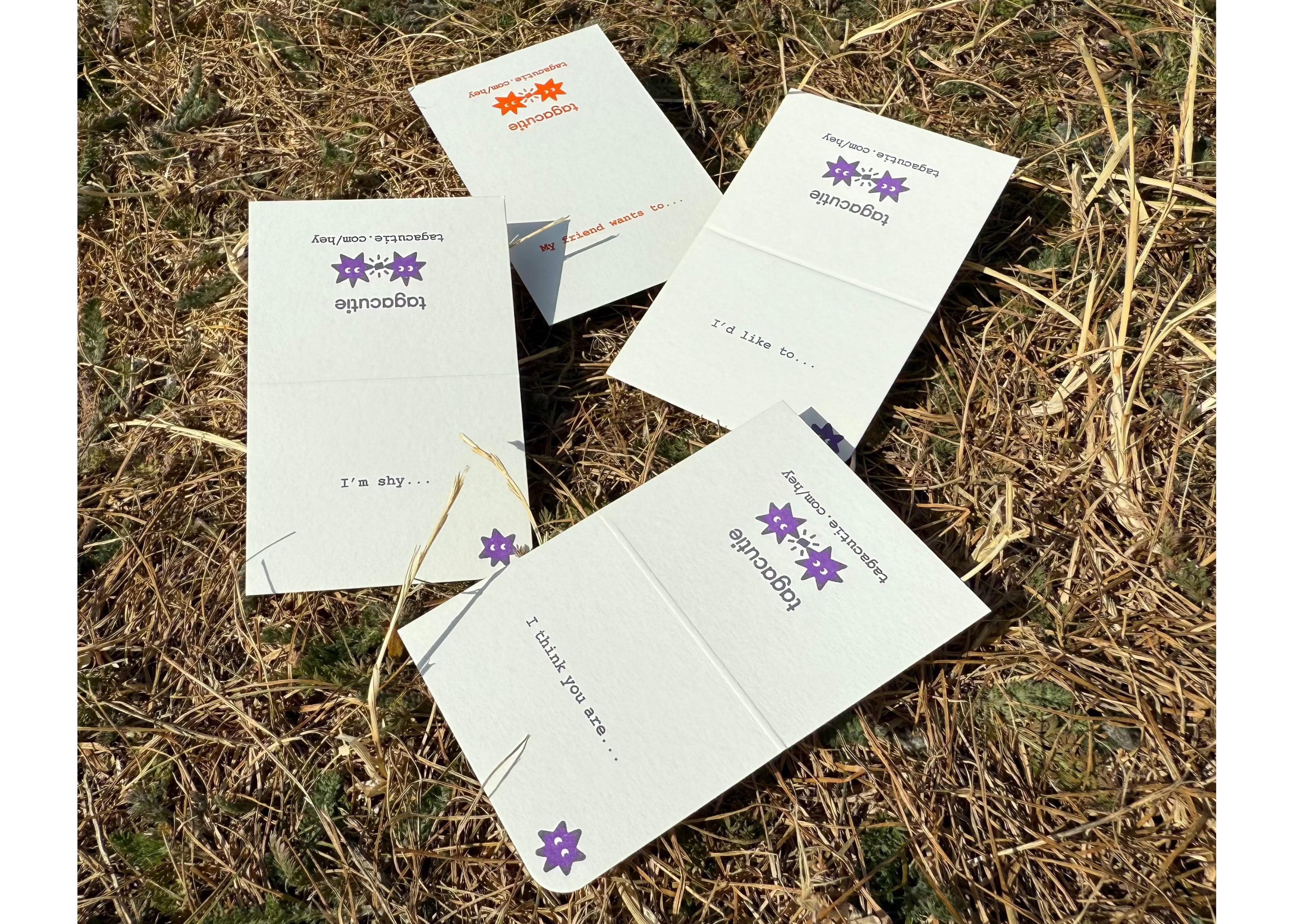 Four white square paper cards or envelopes with purple floral graphics and printed text, scattered on dry brown grass outdoors.
