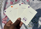A hand holds four small cream-colored cards or envelopes fanned out against a floral fabric background. Each card has a small purple flower-like logo and black text, including the visible brand name 'tagacutie' and website 'tagacutie.