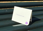 A small white folded greeting card with printed text and a purple flower illustration, placed on a dark green metal bench outdoors.