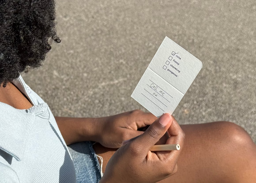 A person sitting outdoors on a bench, holding a small white card or ticket in their hands. The person has dark curly hair, is wearing a light blue ribbed top and denim shorts, and is photographed from an over-the-shoulder angle against an asphalt background.