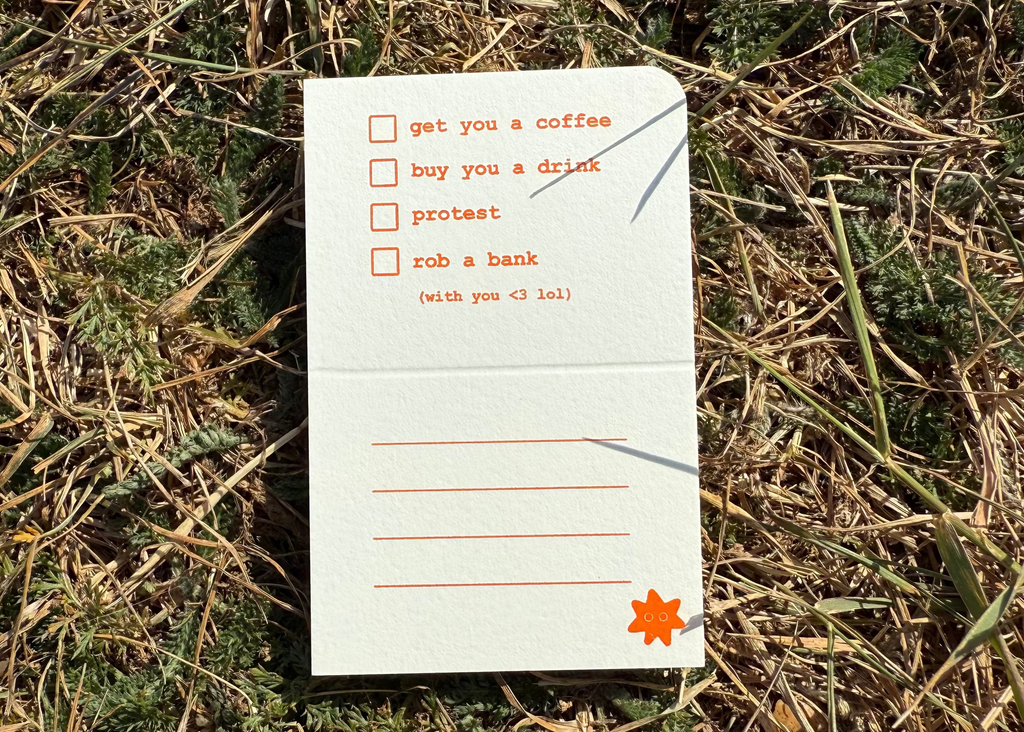 A small white folded paper card or notepad placed on dry grass and green ground. The card has orange printed text and checkboxes, with one checked box containing the phrase 'get you a coffee' and a small orange star near the bottom right corner.