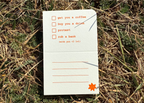 A small white folded paper card or notepad placed on dry grass and green ground. The card has orange printed text and checkboxes, with one checked box containing the phrase 'get you a coffee' and a small orange star near the bottom right corner.